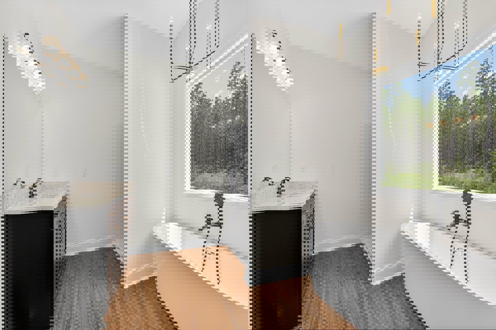 Freestanding white bathtub with exposed black plumbing, wood vanity topped with black rectangular vessel sink, large window framing leafy trees, white lamp shade and ceiling light