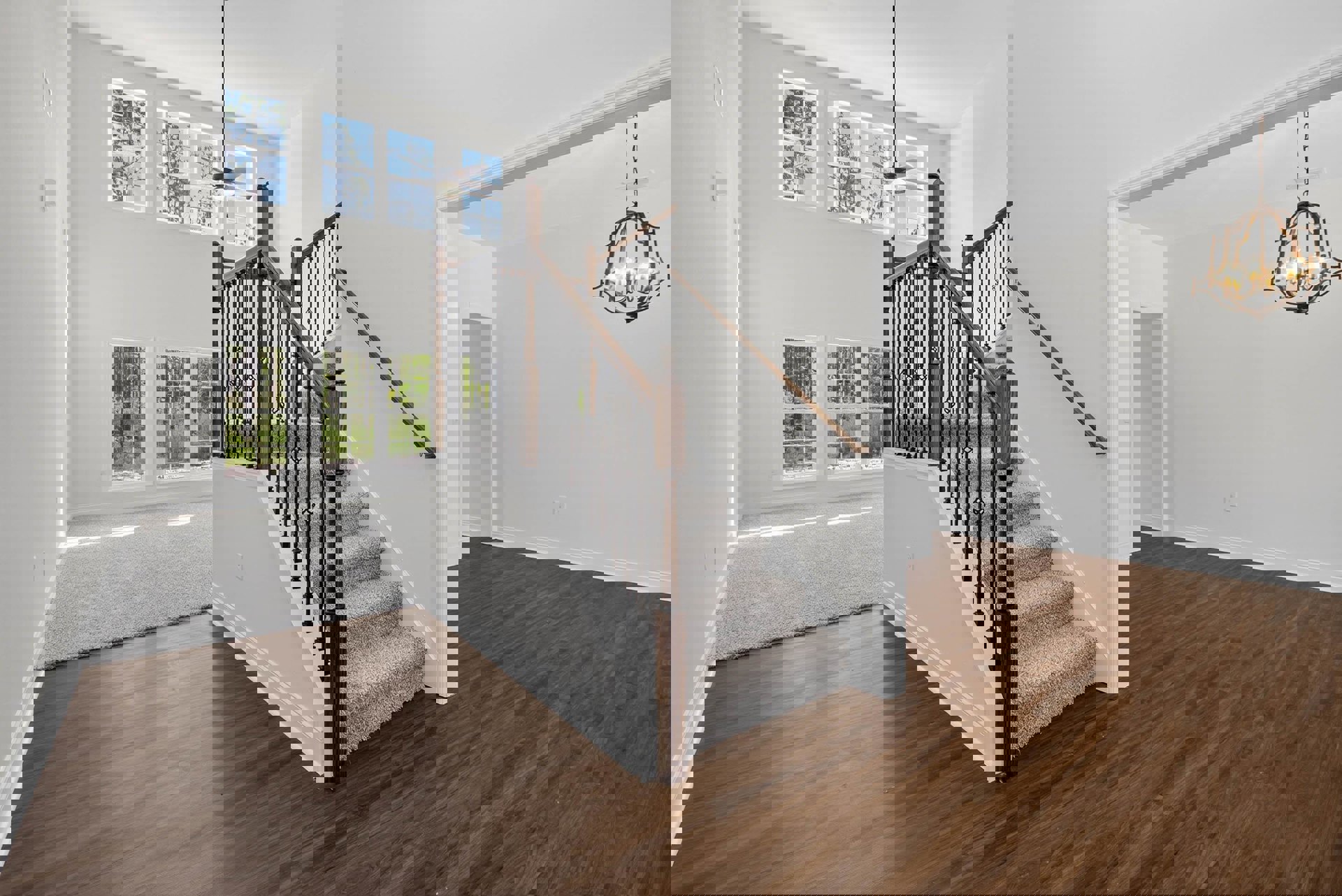 Curved staircase with metal railing and wooden steps, chandelier overhead, large window overlooking trees, white carpeted area adjacent to hardwood flooring