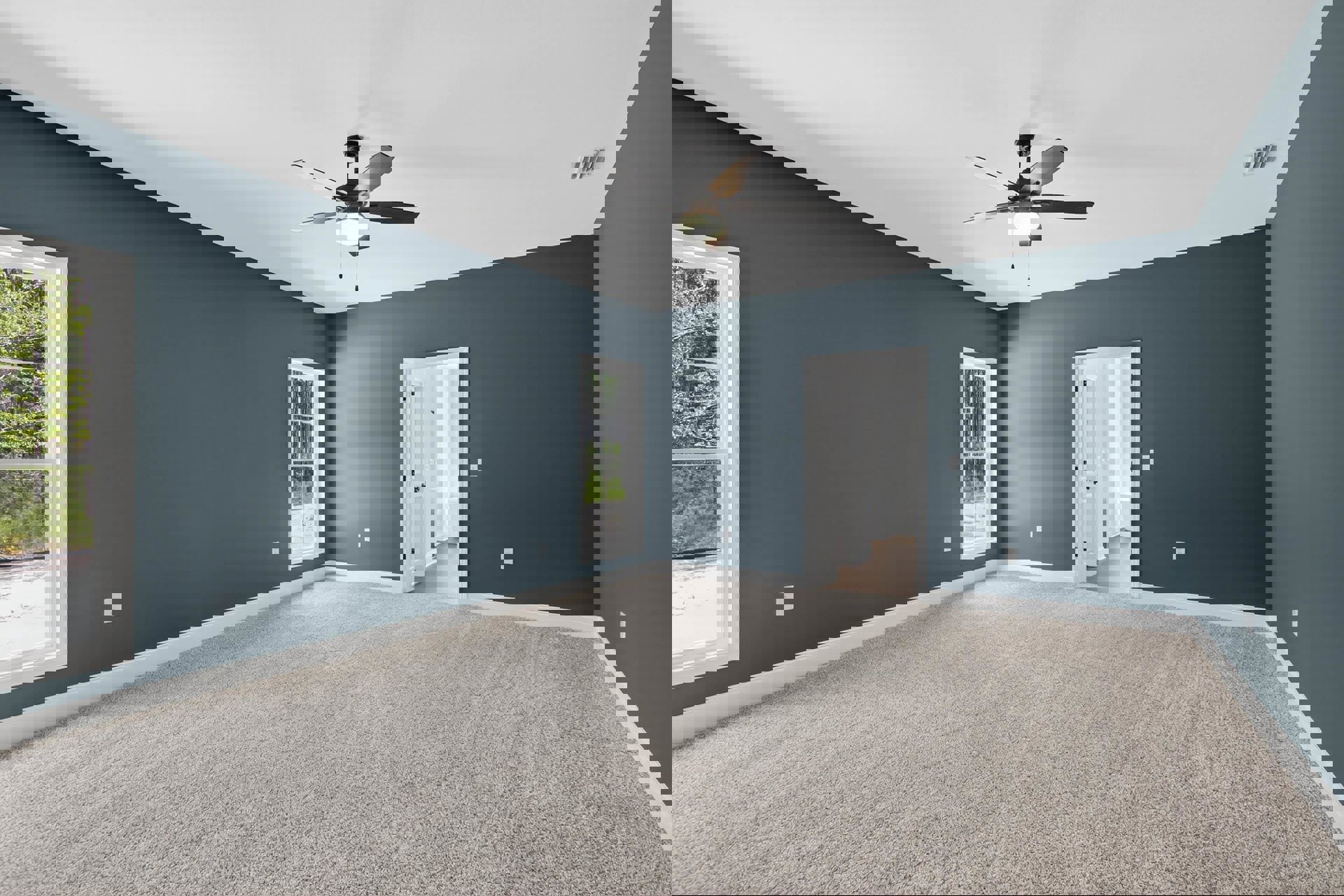 Bedroom with beige carpet, white walls, ceiling fan with light, single window framed in white showing trees outside, and a closed door.