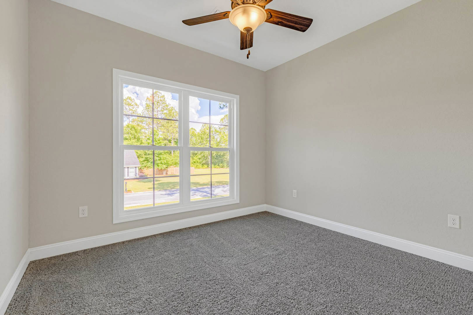 Bedroom with grey carpet, white walls, ceiling fan with light fixture, large window overlooking trees, white electrical outlet on wall