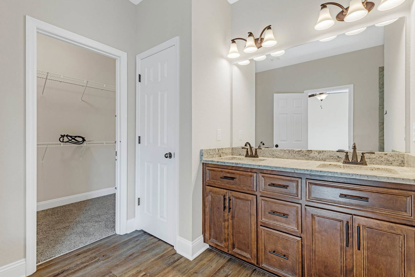 Bathroom with double sink vanity, wide mirror above, white cabinetry, silver faucets, tiled backsplash, white door with silver knob, ceiling light fixture, open drawer, wire shelf