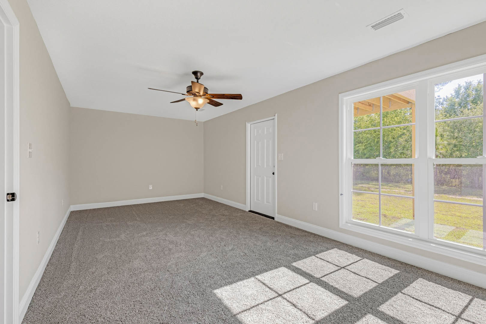 Carpeted bedroom with white walls, ceiling fan with light, large window showing outdoor greenery, and white door with black hardware