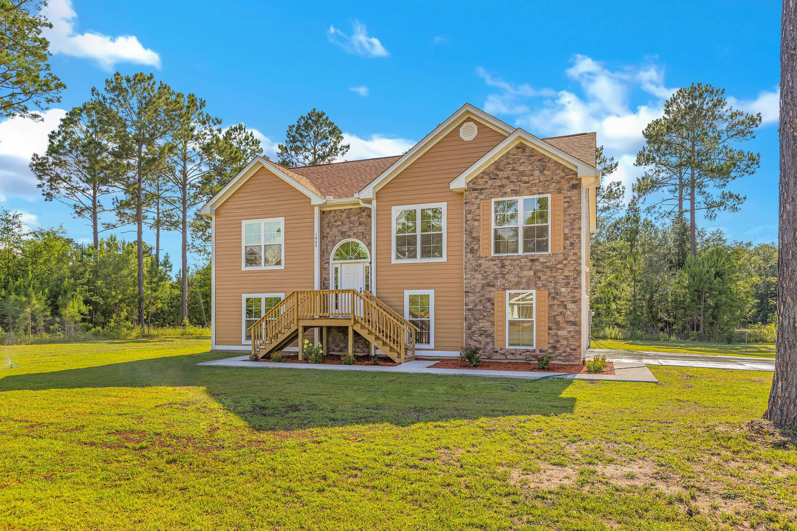Two-story home with white-framed windows, wood deck and railing, green lawn, mature trees, and partly cloudy sky in the background
