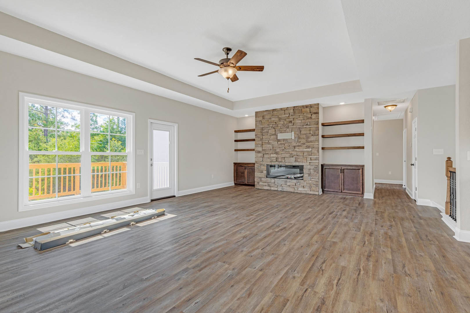 Spacious living room featuring hardwood floors, stone fireplace with rectangular window, ceiling fan with light fixture, built-in cabinet, and large window overlooking trees