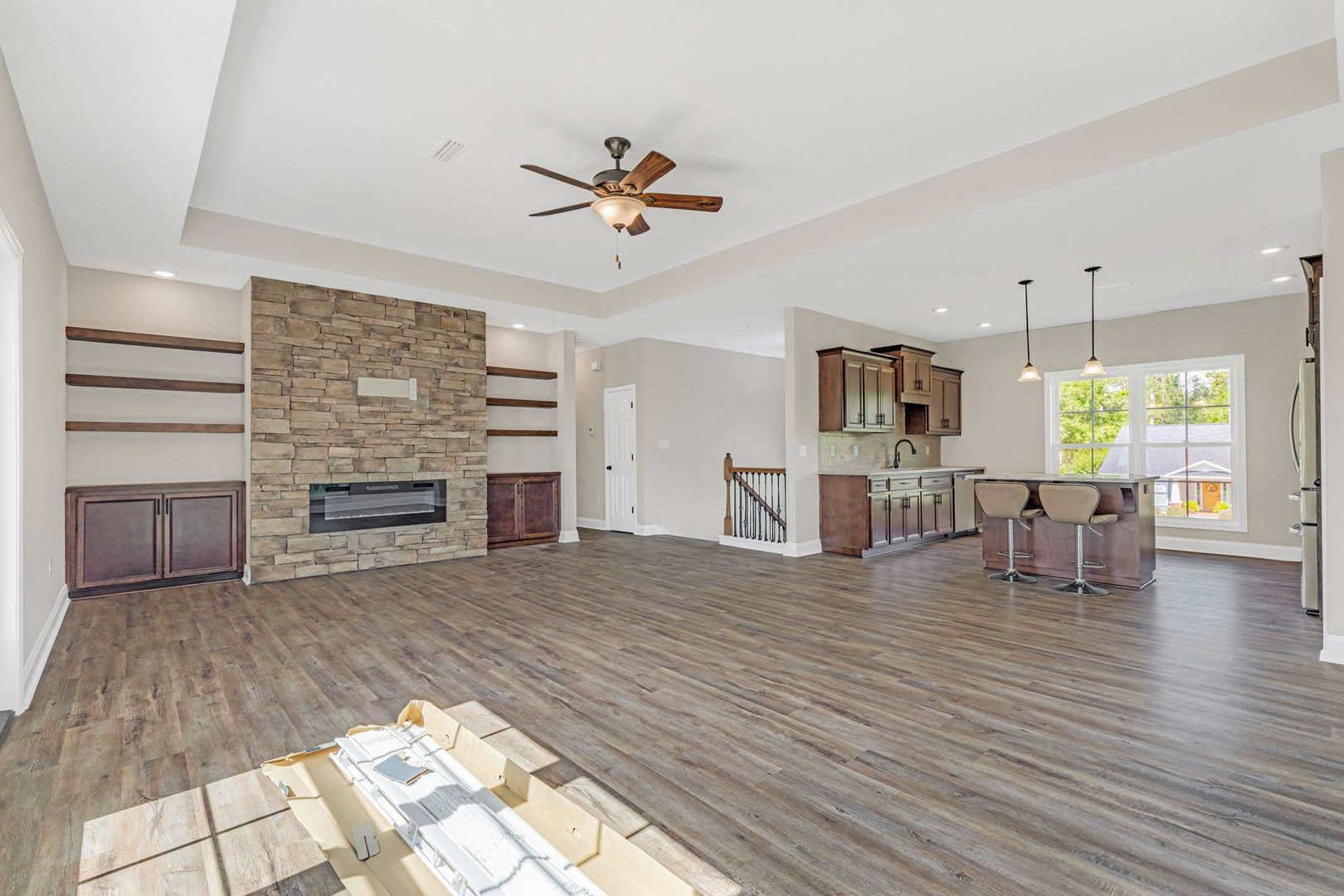 Spacious living room featuring hardwood floors, stone fireplace with white mantel, ceiling fan with light fixture, neutral walls, and two upholstered chairs beside a kitchen