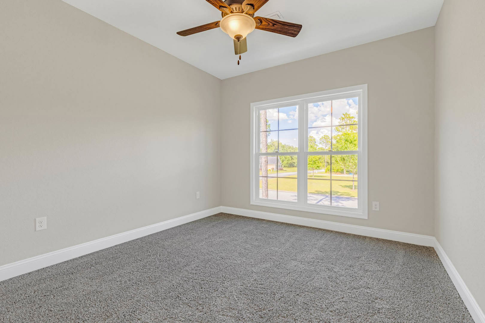 Carpeted room with white walls, ceiling fan with light fixture, large window showing trees outside, white electrical outlet on wall, crown molding along ceiling.