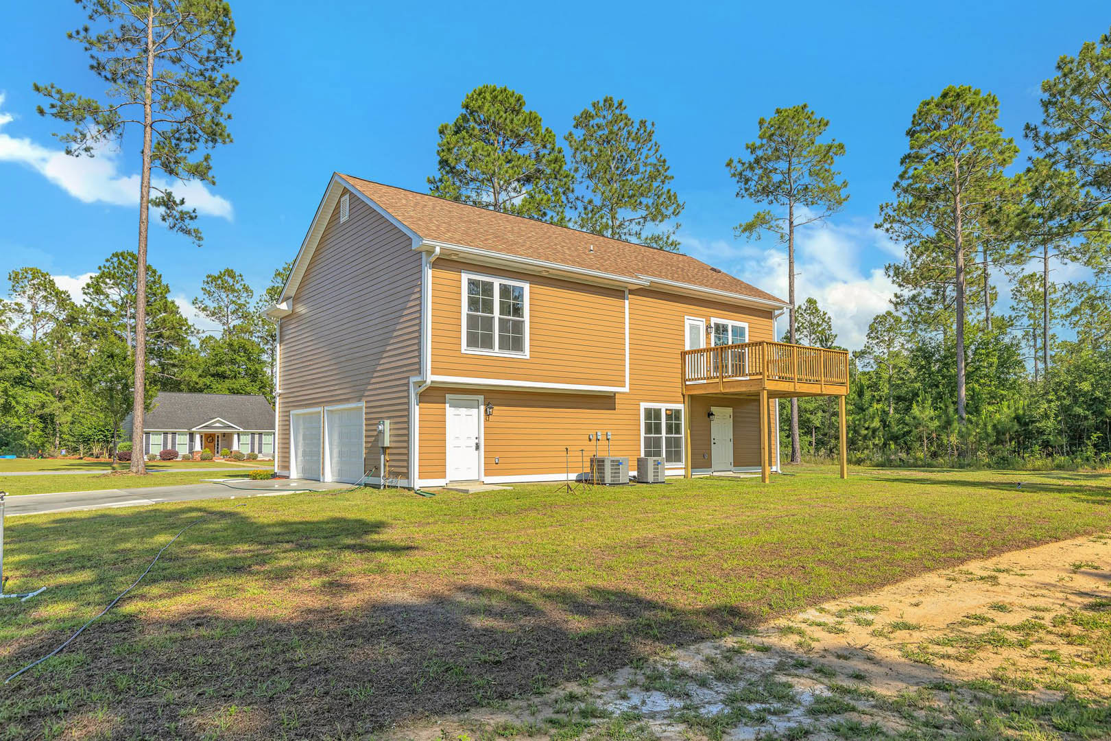 Wood deck overlooking a green yard, white cottage-style house with large windows, tall trees with dense foliage in the background, blue sky with scattered clouds