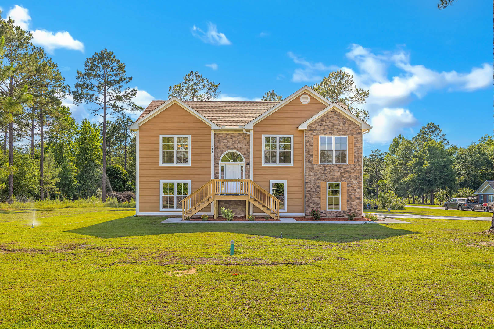Two-story house with white-trimmed windows, wooden deck and railing, front staircase, green lawn, tall trees and cloudy sky in background