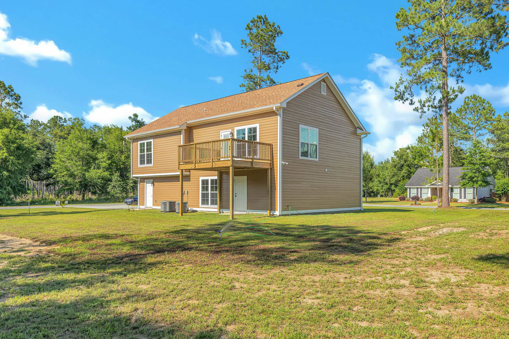 Two-story house with covered front porch, manicured lawn, sprinkler watering grass, large windows, balcony, and tall trees in background under partly cloudy sky