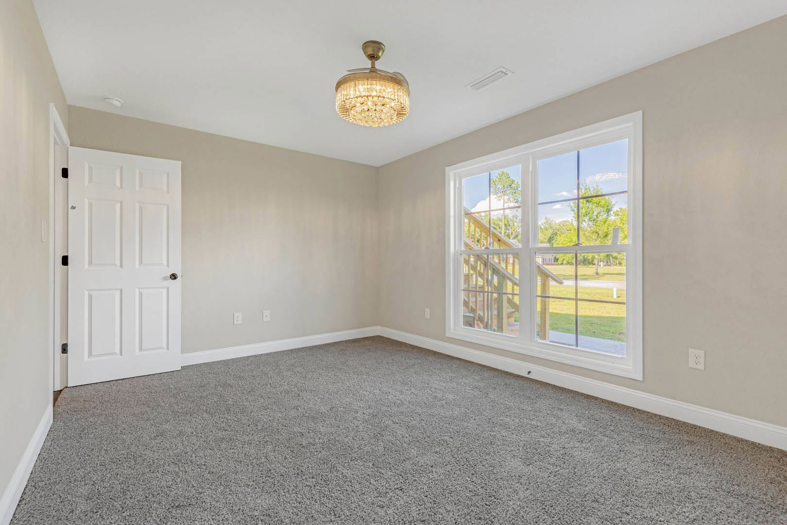 Chandelier hanging from white ceiling, large window with backyard view, white door with silver handle, light-colored carpet flooring, neutral walls with crown molding