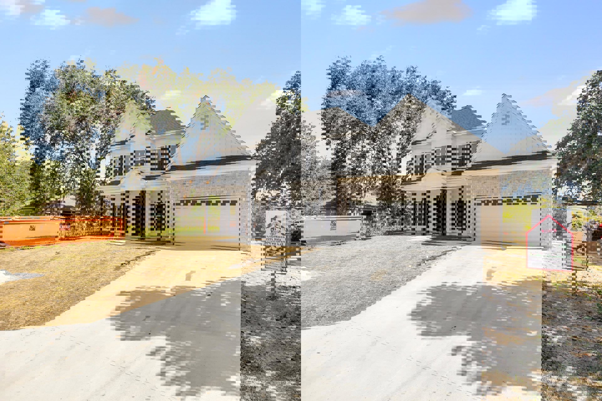 Two-story house with light siding, attached garage, paved driveway, green lawn, mature trees, large orange dumpster near the curb, cloudy sky overhead