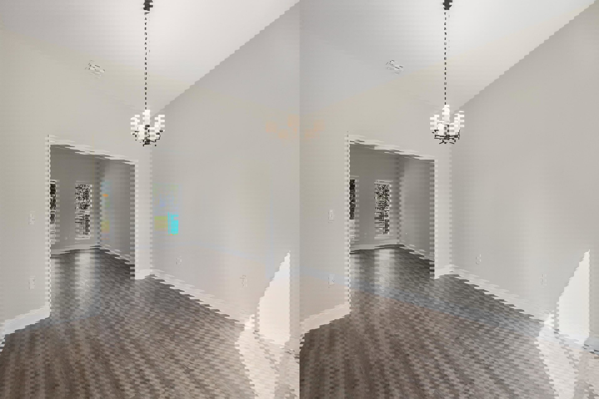 Wood flooring in a spacious room with a white plaster wall, ceiling vent, and a chandelier featuring white lamps; large window offers a view of a green container outside.