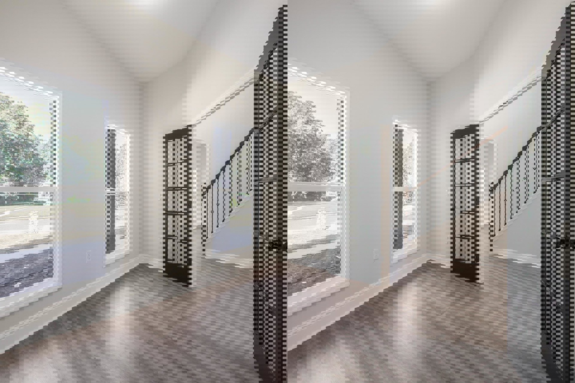 White oak wood flooring in a bright room featuring a glass-paneled door, large window, and staircase with black metal railings; white brick accent wall visible near entry.