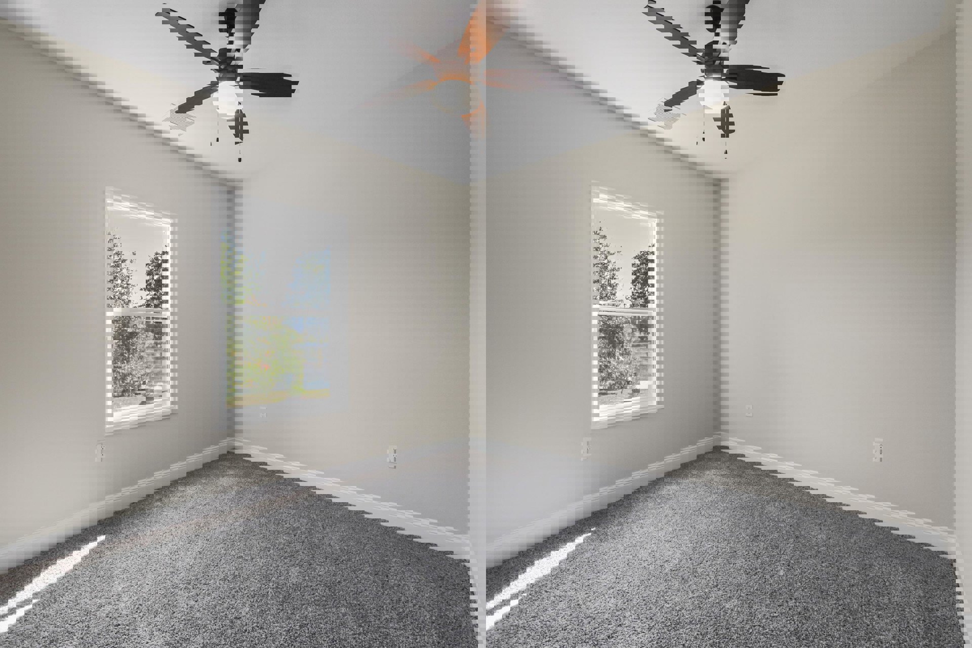 Ceiling fan with integrated light fixture above grey carpeted floor, large window revealing green trees outside, white plaster walls