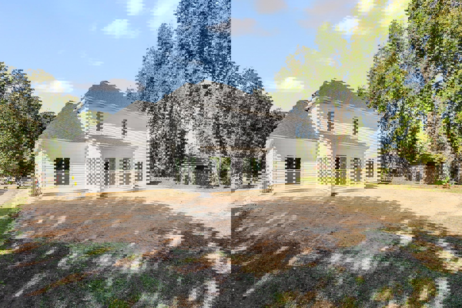 White house with gray roof, dirt yard in foreground, several trees behind, gray air conditioning units near side wall, large windows visible