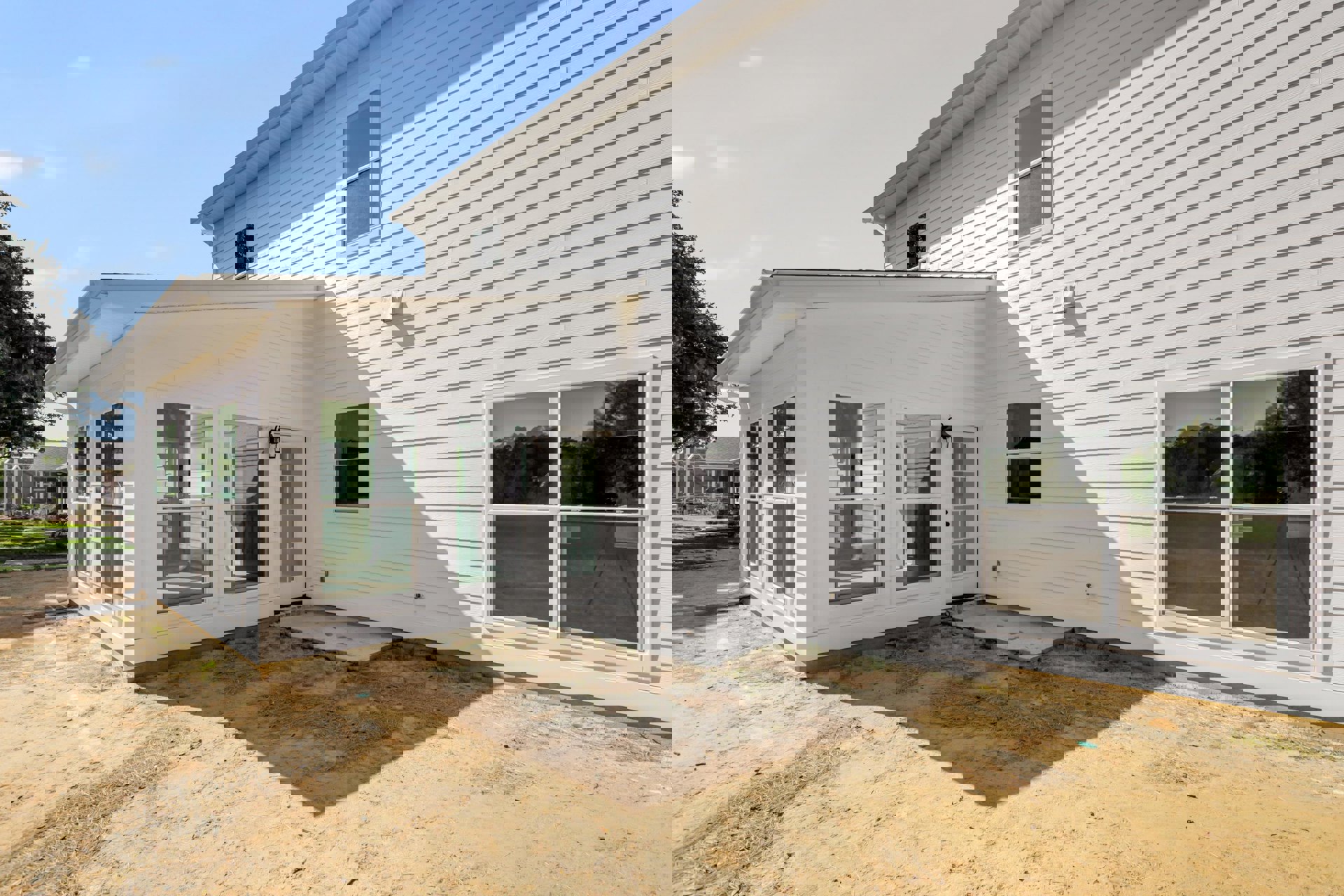 Modern home exterior with white siding, large window reflecting trees, patio area with dirt ground, white framed door, and minimalistic white wall.