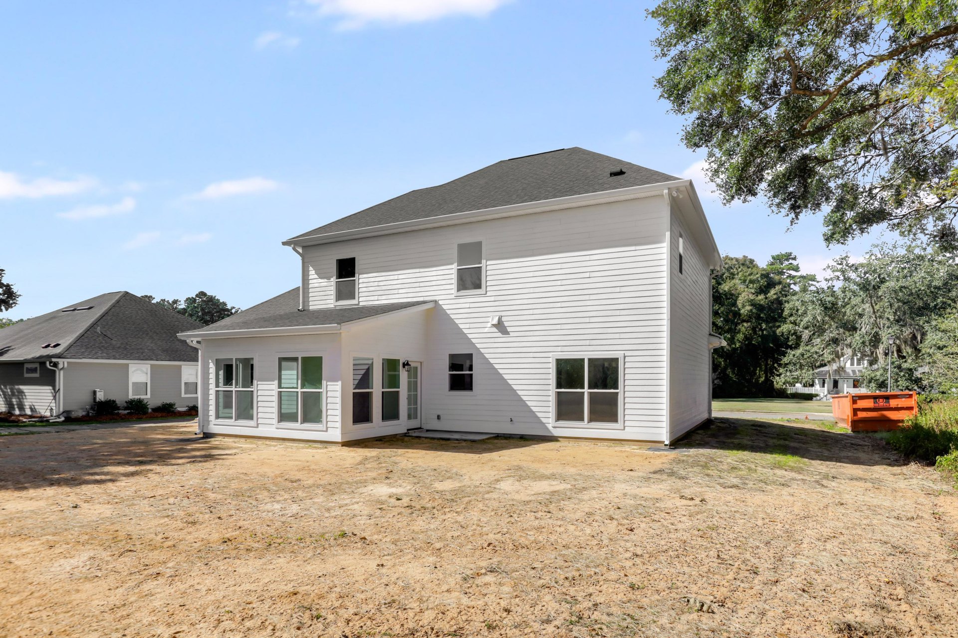 White house with black roof, large glass windows with white frames, dirt backyard, small porch, trees and clouds in the background, dumpster near the house.
