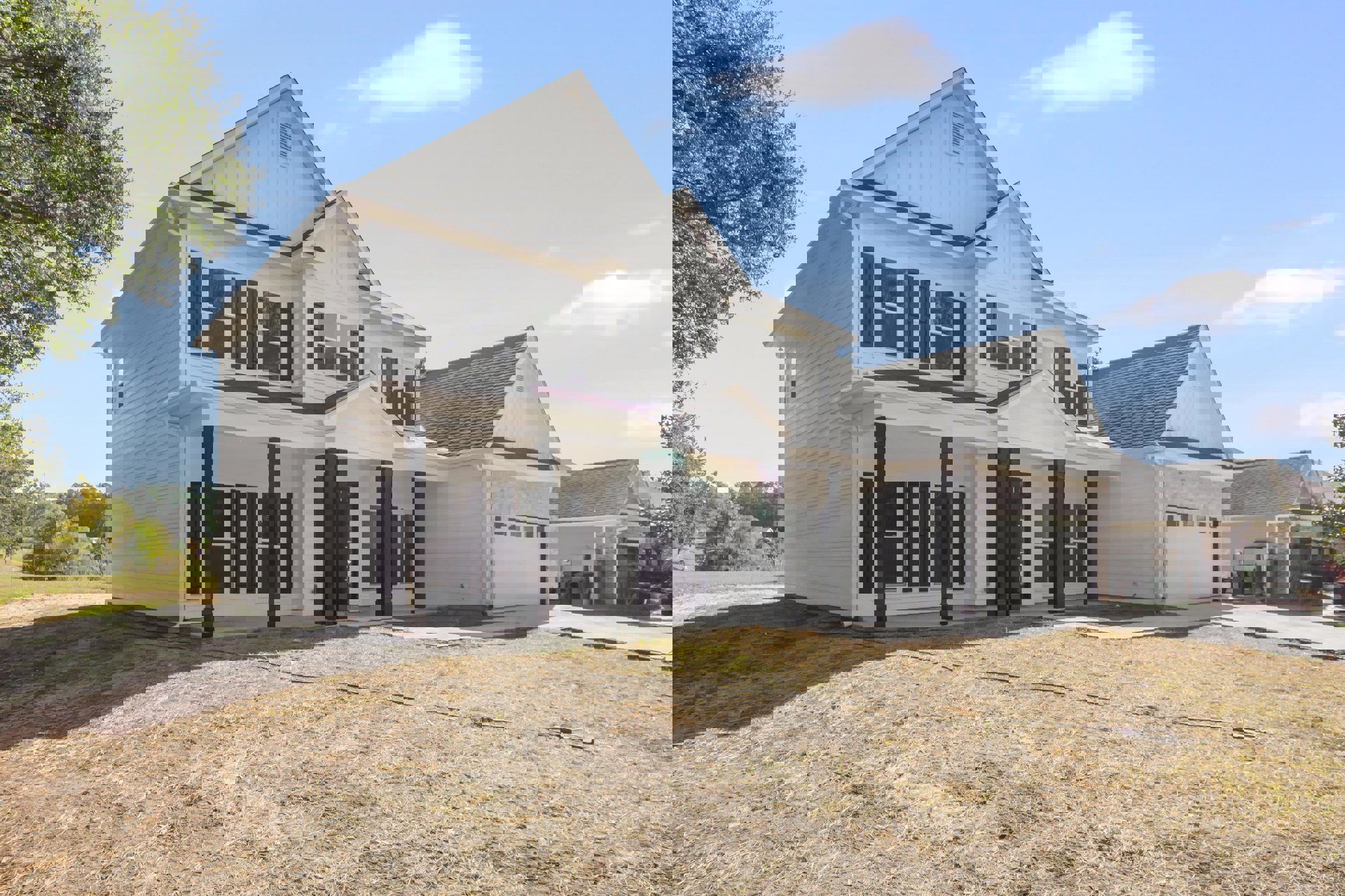 White two-story house with black columns, wide driveway, manicured lawn, garden hose on grass, mature trees, and blue sky with scattered clouds