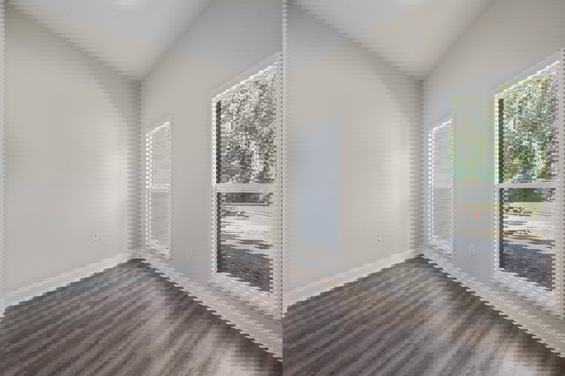 Sunlit room featuring wide plank wood flooring, large windows with views of trees and a road, white plaster walls, and minimalist interior design.