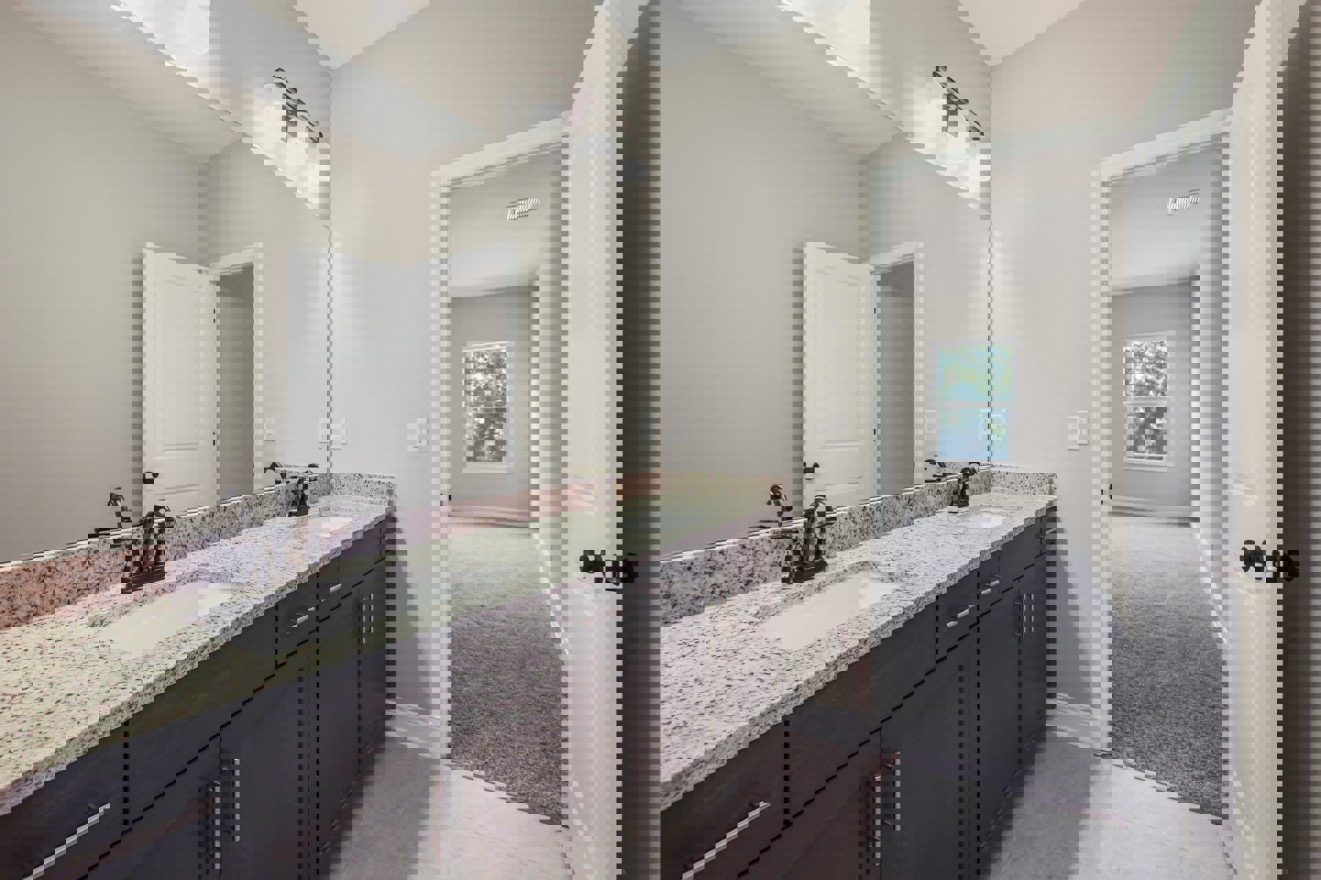 Bathroom with granite double vanity, large mirror, tile flooring, white cabinetry, chrome faucets, window overlooking trees, open white door leading to carpeted hallway