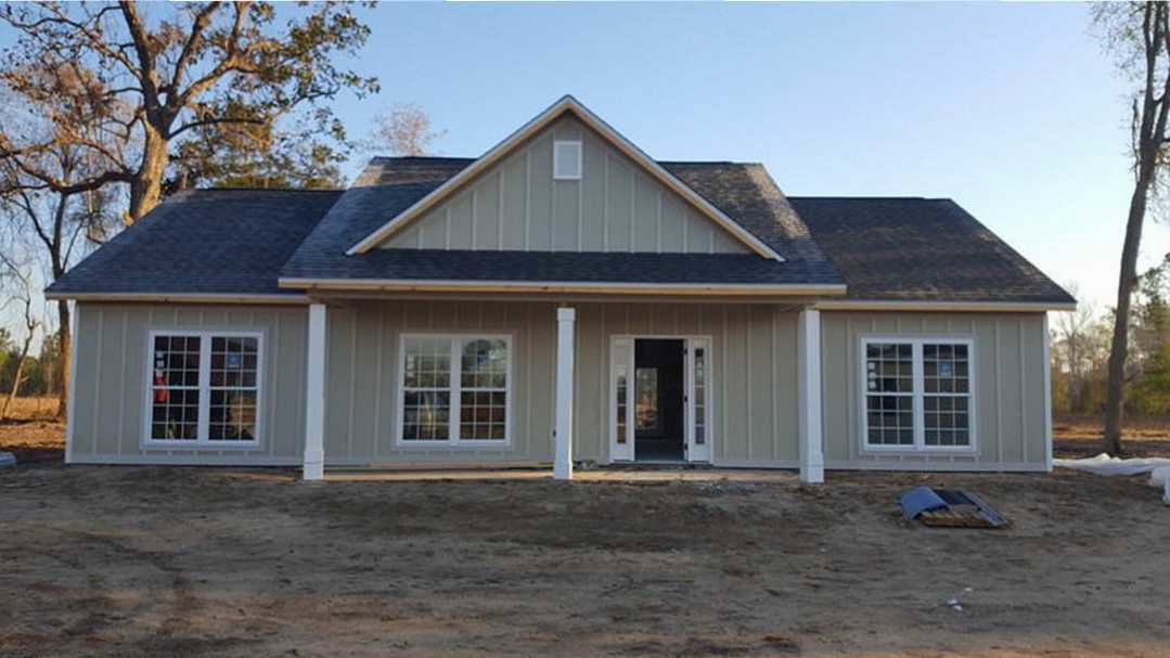 Two-story house under construction with gray siding, black front door with glass panes, white columns, multi-pane windows, and dirt yard in foreground