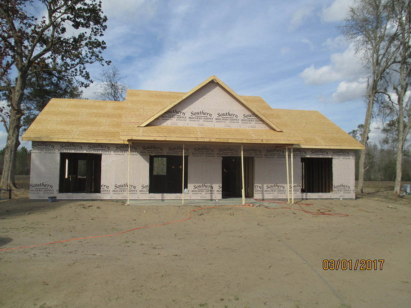 Partially built house with exposed plywood walls, installed roof, several windows, construction materials scattered on dirt ground, and nearby tree under cloudy sky
