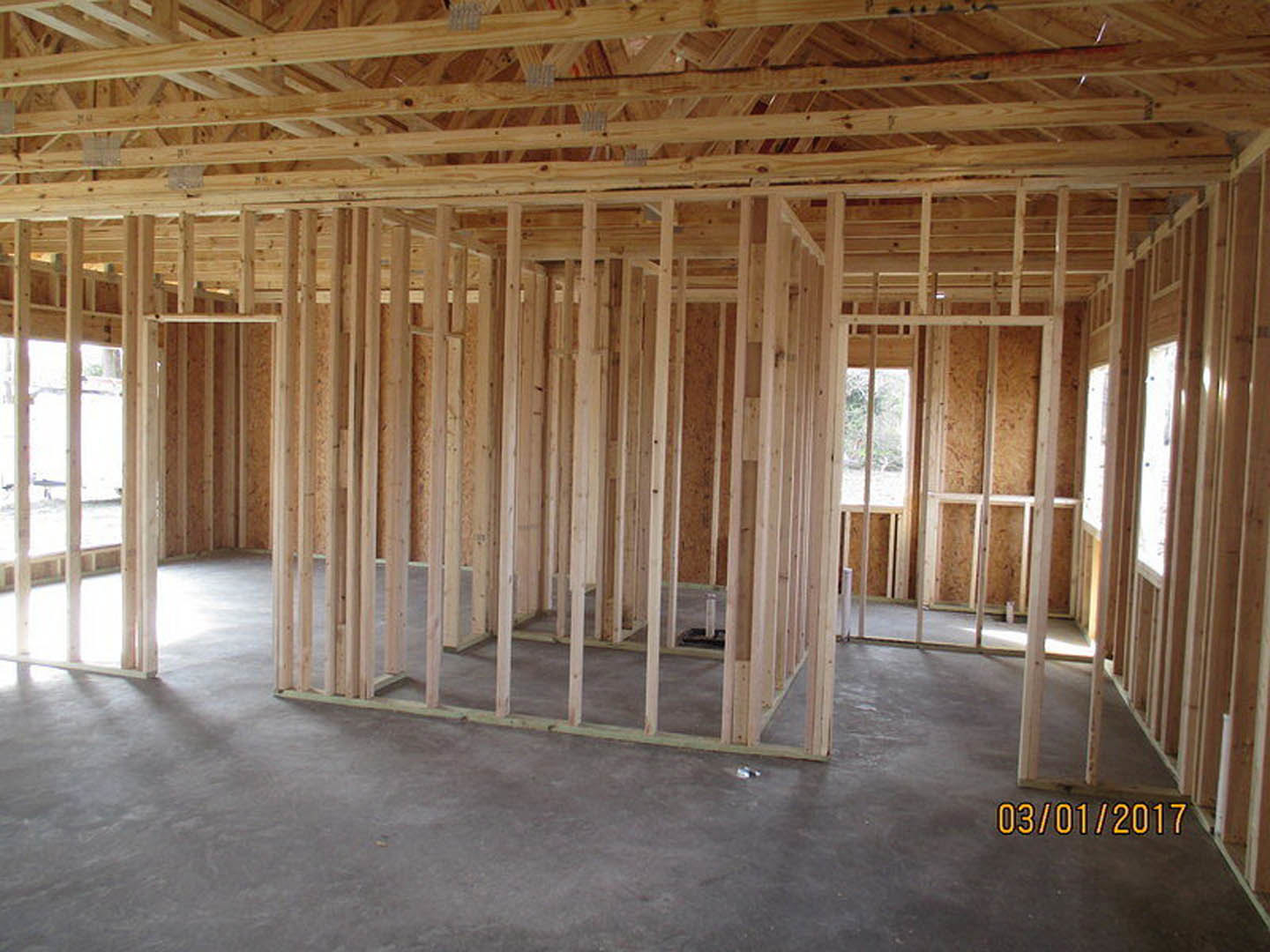 Exposed wood framing with thick beams, unfinished concrete floor, large window showing bright sky, open ceiling with visible insulation