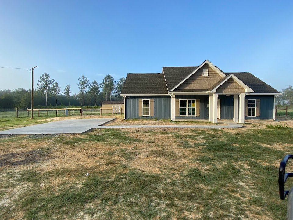 Two-story house with white trim windows, gray roof, and fenced grassy yard, concrete walkway leading to covered porch, blue sky and trees in background