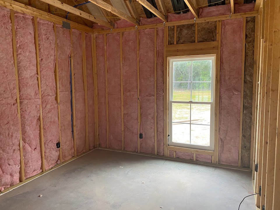 Concrete floor partially covered by pink cloth, exposed wooden ceiling beams, white-framed window overlooking grassy field, black wire protruding from wall