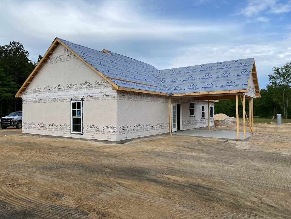 Partially built house with exposed plywood roof marked by blue tape, construction truck parked nearby, window displaying permit sign, dirt driveway leading to site, mature trees in