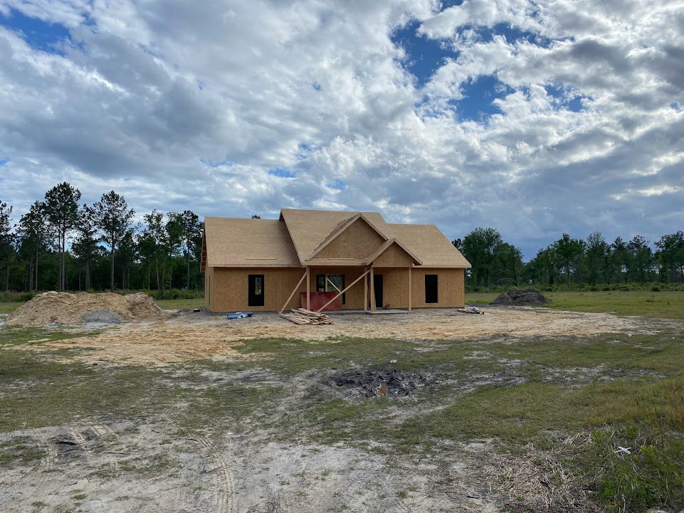Wood-framed house under construction on a grassy lot, exposed foundation and dirt mound in foreground, blue sky with scattered clouds, trees lining the background