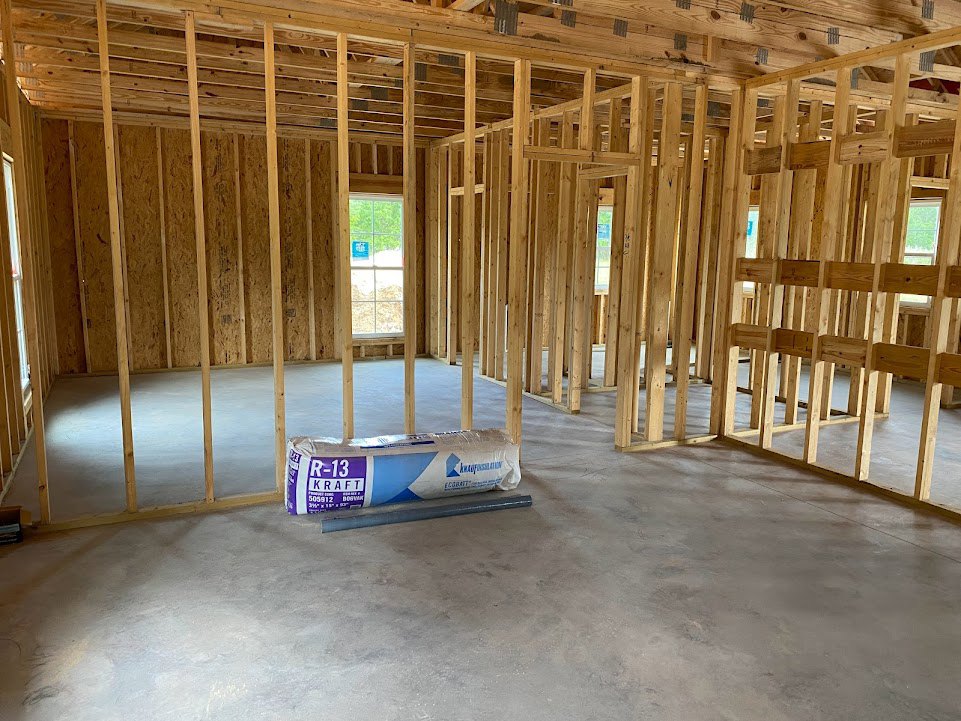 Unfinished room with exposed wood framing, concrete floor, large cement bag, window with blue sign, and a chair