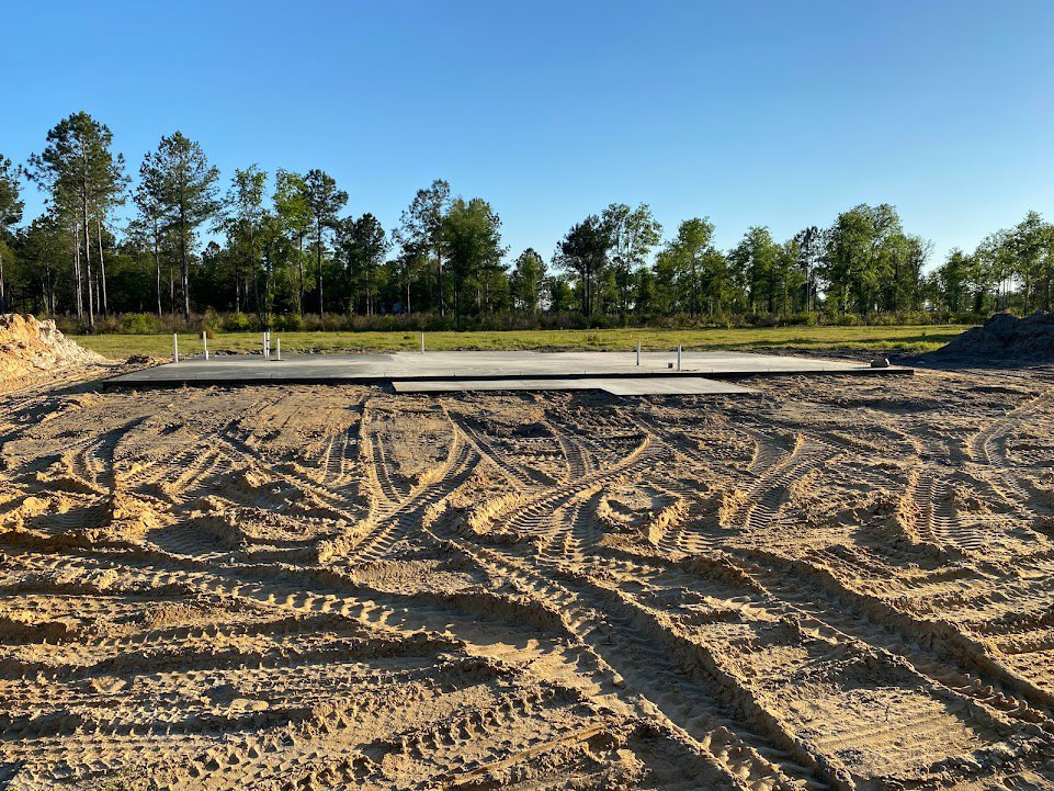 Sandy driveway with visible tire tracks leading toward grassy field and cluster of trees under clear blue sky