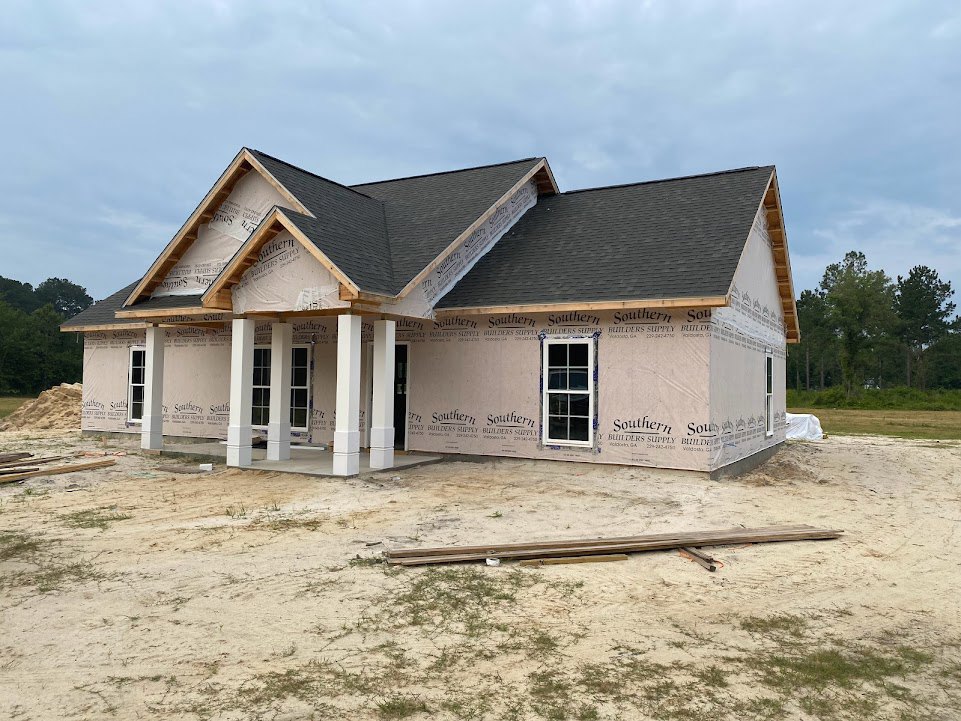 Partially built house with exposed wooden framing, white window frame, roof with construction sign, scattered wood piles on dirt ground, trees in background