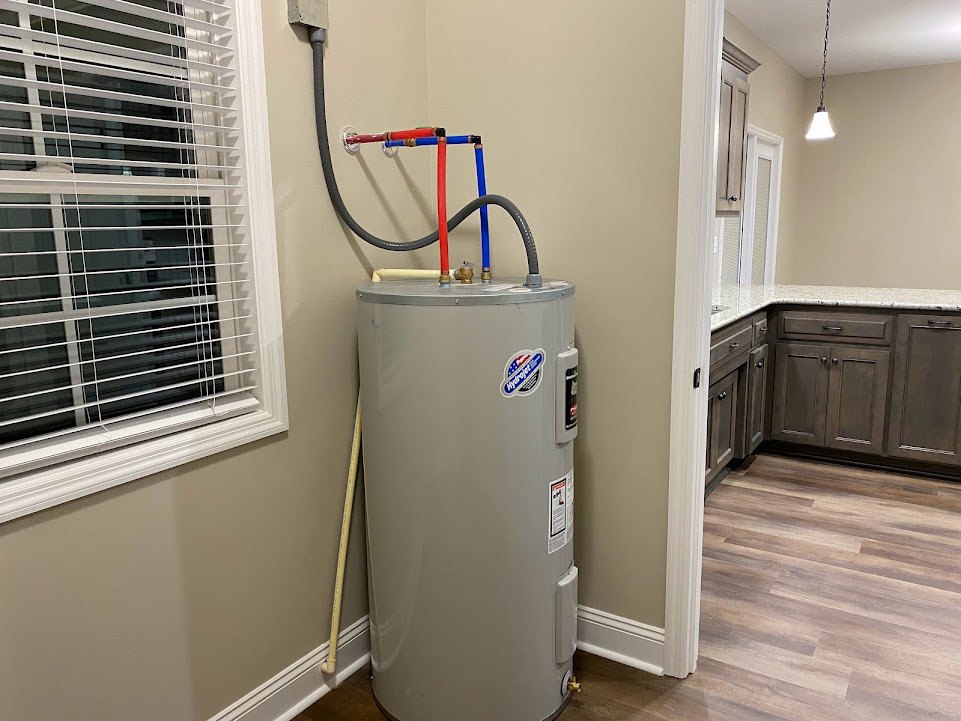 White water heater with attached pipes and manufacturer sticker, positioned in a corner near a window with closed blinds, adjacent to a kitchen countertop and refrigerator on a