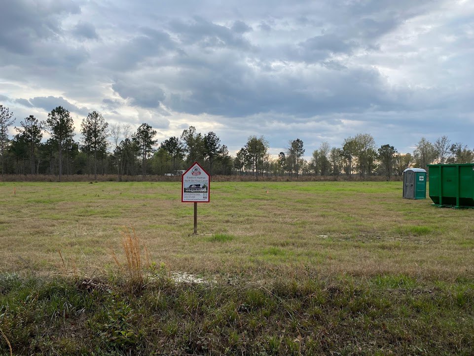 Wooden post with a house-shaped sign in grassy field, green dumpster and portable toilet nearby, trees and clouds in background