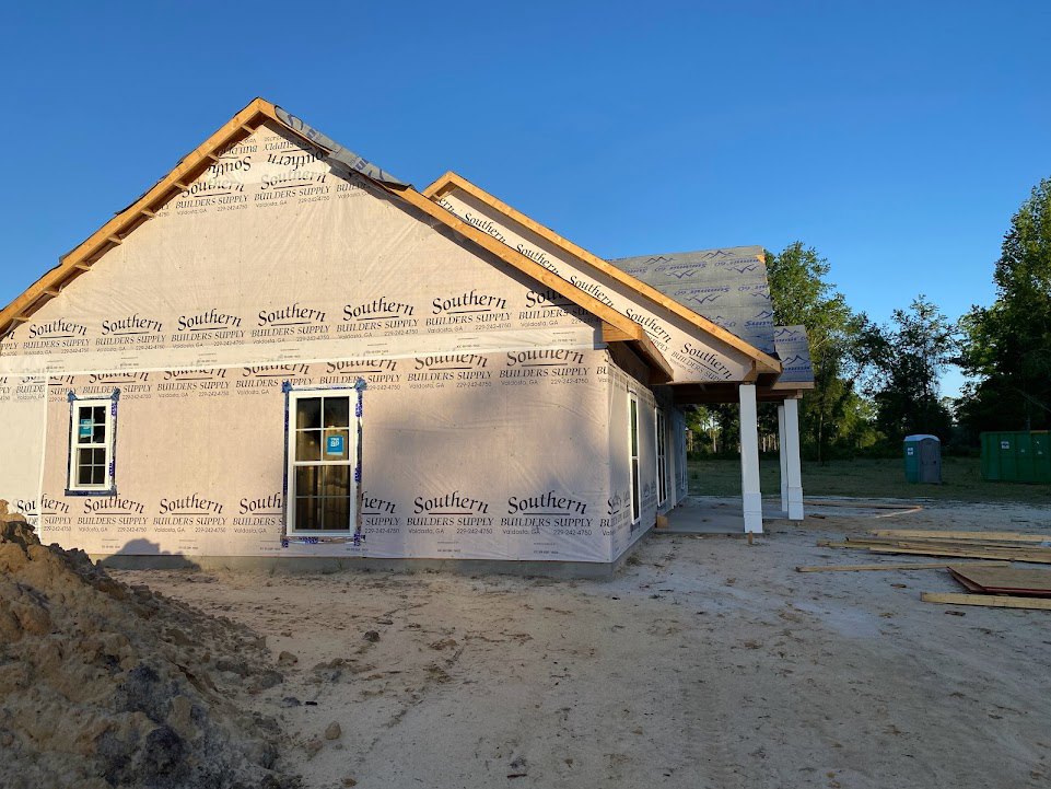Partially built house with exposed framing, plastic sheeting, and a blue sign in the window; pile of dirt and green bin in front; mature trees surrounding construction site.