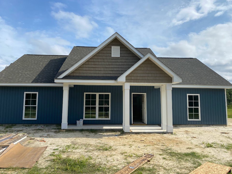 Partially built house with blue siding and white trim, white-framed windows and door, person standing in window opening, brown construction paper on ground, clear blue sky overhead