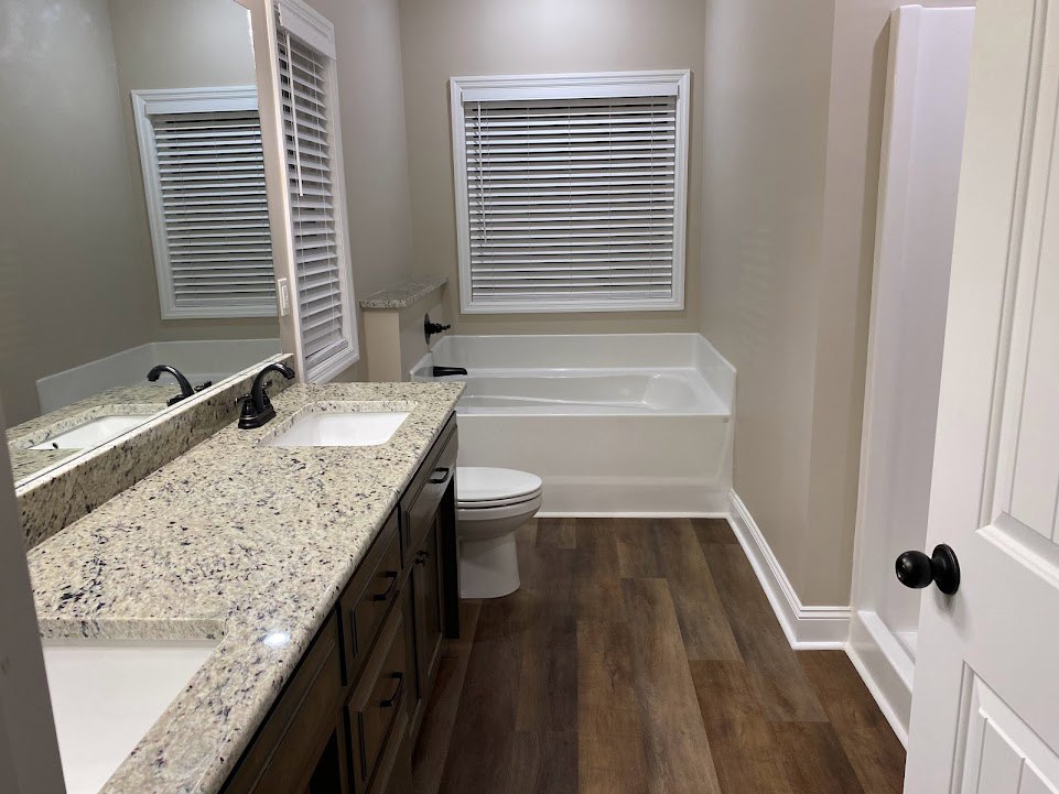 Bathroom with marble countertop, undermount sink, black faucet, white walls with black door knob, window with blinds, close-up of toilet, wood flooring with white trim