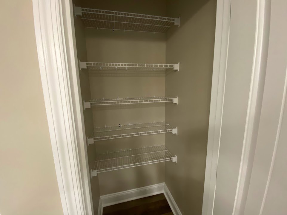 Walk-in closet with white wire shelving mounted on plaster walls, white metal rack attached to ceiling, and rectangular shelf with square pattern.
