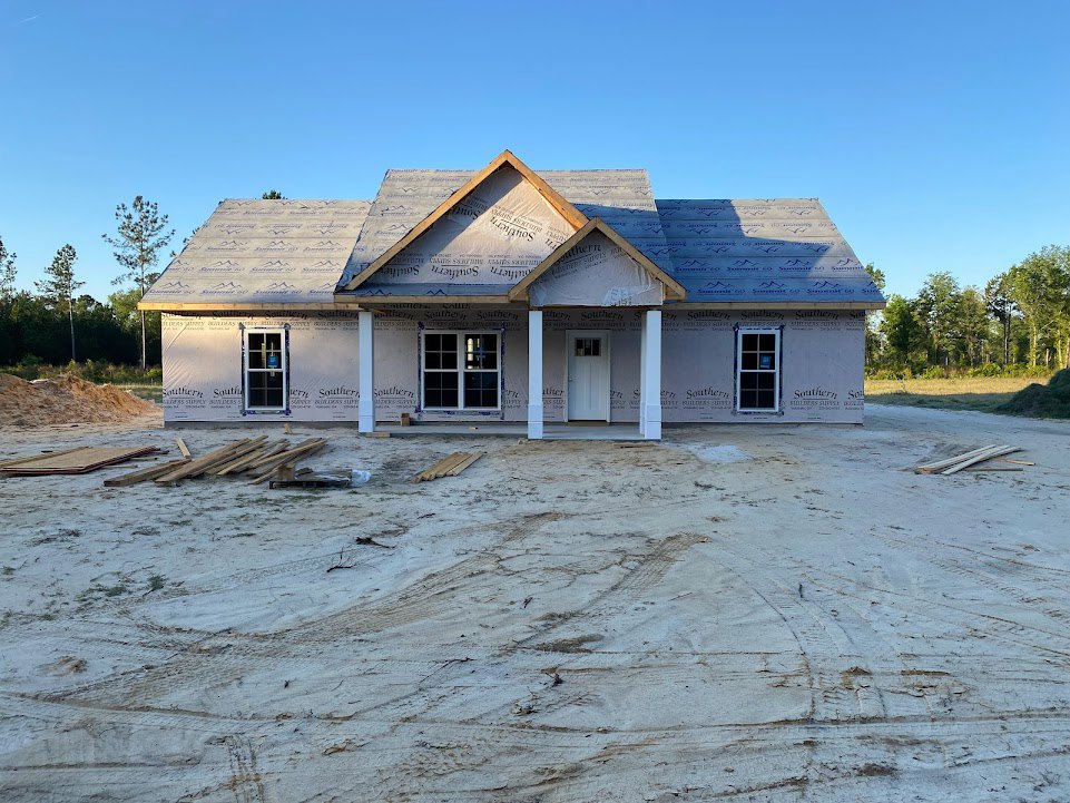 Partially built house with multi-pane windows, white columns, and triangular roof, surrounded by dirt field and forested trees under open sky
