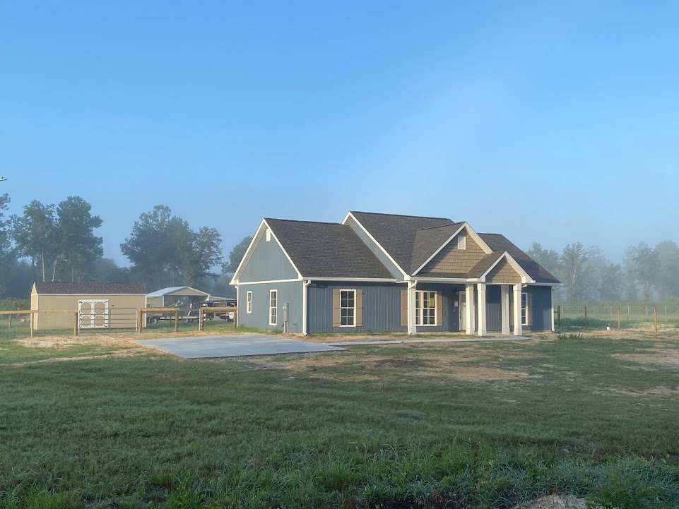 Two-story house with blue roof, white siding, and white front door, set on a grassy lawn with a wooden fence and trees in the background under a clear blue sky