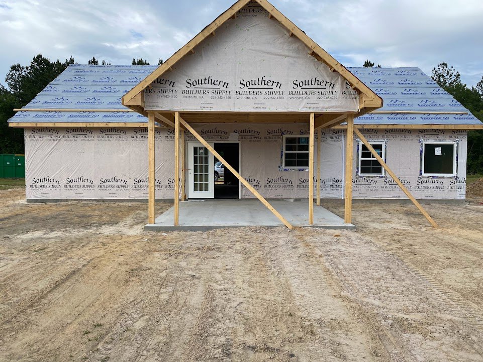 Wood-framed house under construction with exposed beams, partially installed roof, white door with glass panes, and window openings; exterior siding not yet finished.