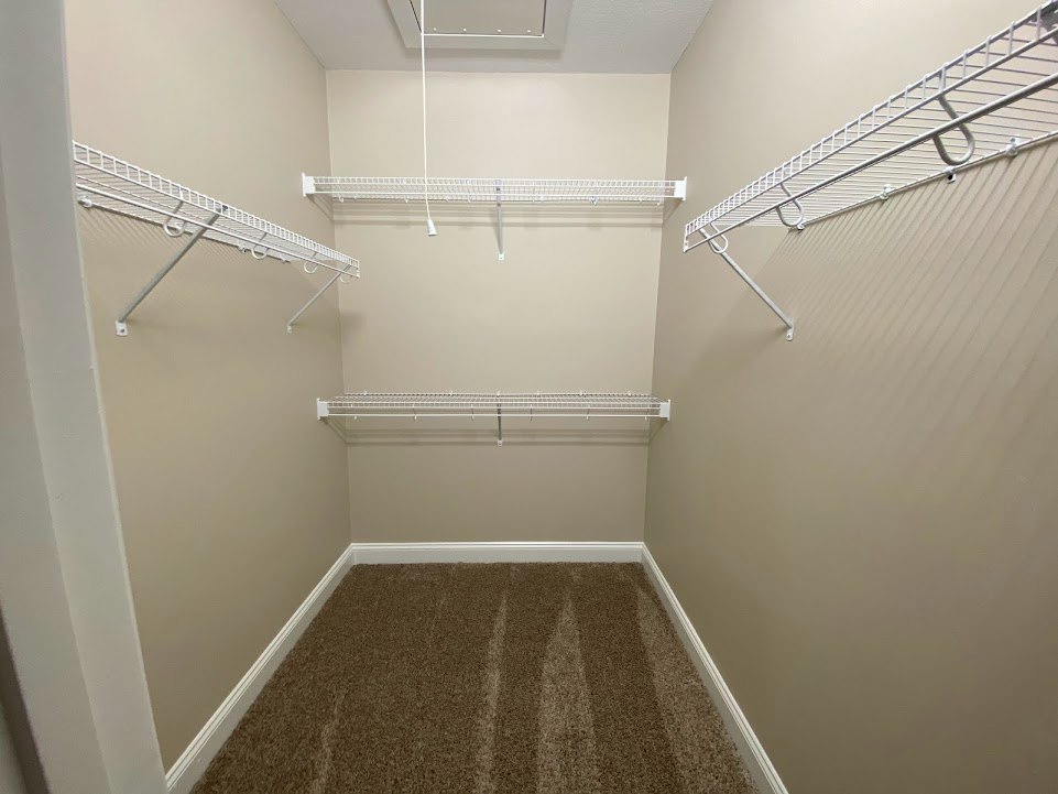 Walk-in closet with white shelving, metal rods for hanging clothes, and light carpet flooring with a white border