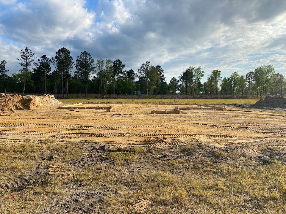 Dirt field with tire tracks, large pile of soil, scattered trees and light pole under cloudy sky in rural grassland