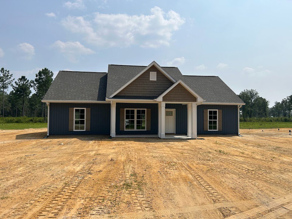Partially built house with white door and window featuring white trim, surrounded by dirt ground with tire tracks, trees in background, cloudy sky overhead