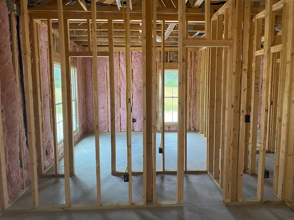 Unfinished room with exposed wood walls marked in black, yellow electrical cable hanging from ceiling, window overlooking green field, construction materials scattered on floor