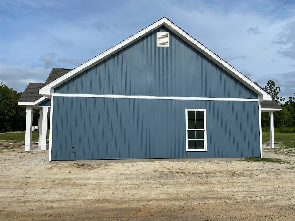 Blue house with white trim and white-framed window, white vent on blue siding, dirt hill beside exterior wall, American Gothic House visible in background, trees and cloudy sky