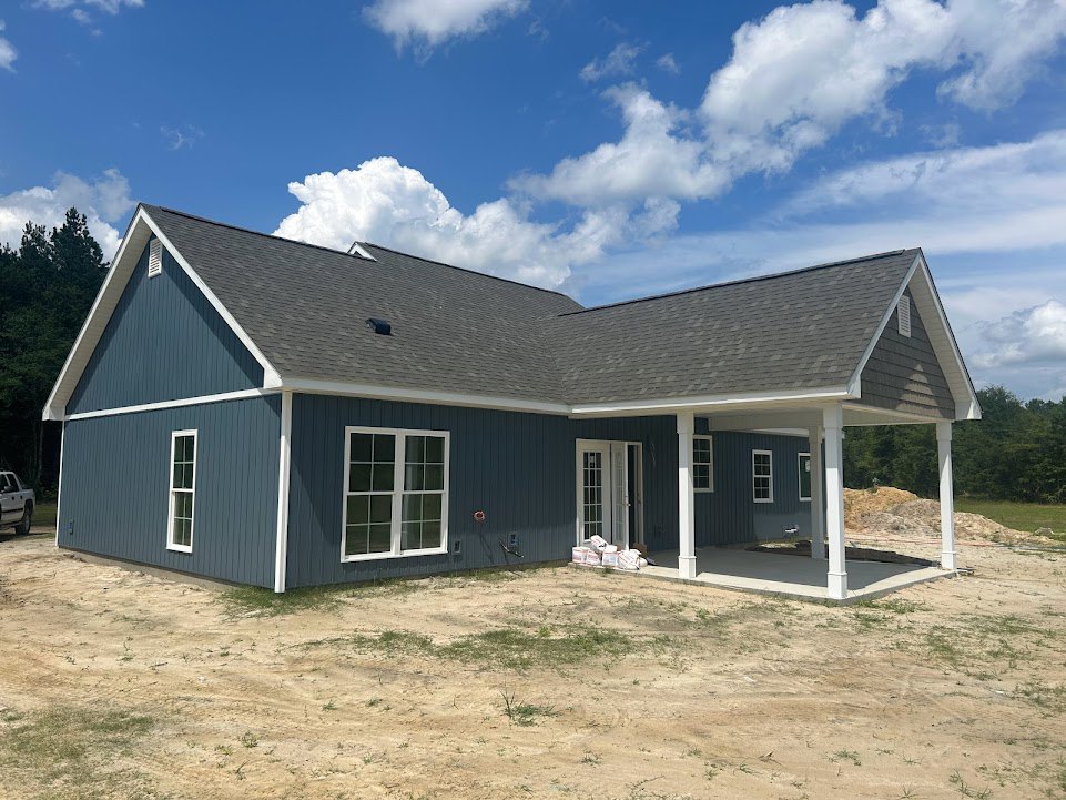 Partially built house with blue exterior walls, white columns, white framed windows, glass paneled door, dirt yard, and cloudy sky overhead