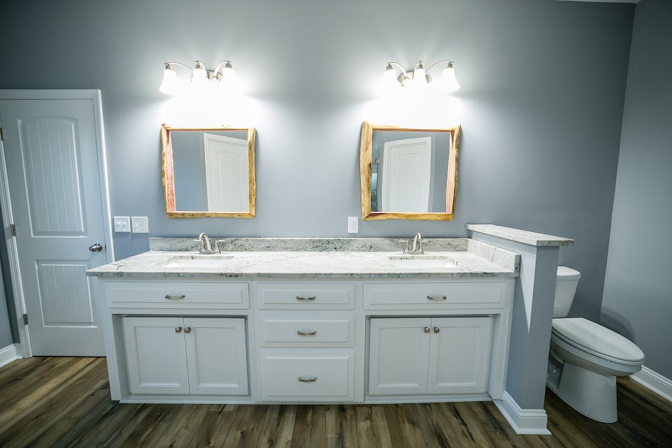Bathroom with white cabinets, marble countertops, two wooden-framed mirrors, silver hardware, white toilet, and white door with silver handle